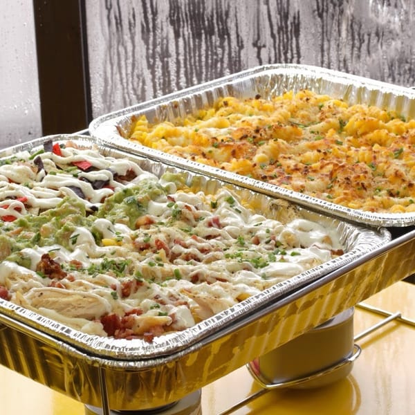 Two foil pans of food, one with mac and cheese, the other with loaded nachos, on a yellow table.