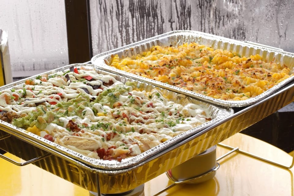Two foil pans of food, one with mac and cheese, the other with loaded nachos, on a yellow table.