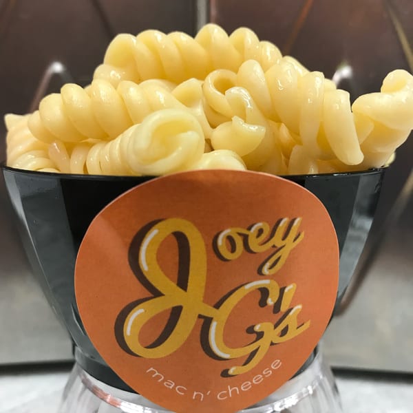 A black cup of Joey G's mac n' cheese, featuring spiral pasta, on a clear stand.