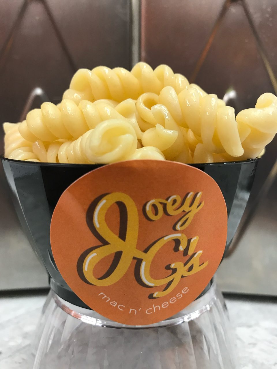 A black cup of Joey G's mac n' cheese, featuring spiral pasta, on a clear stand.