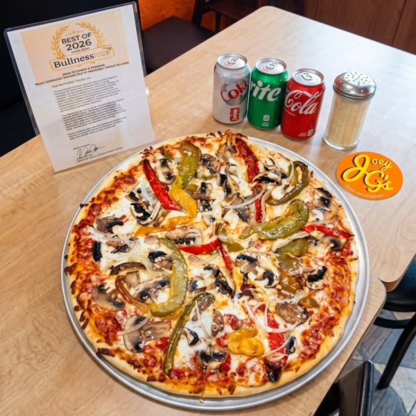 A Joey G's pizza with mushrooms, peppers, and onions, three soda cans, and a spice shaker on a wooden table.