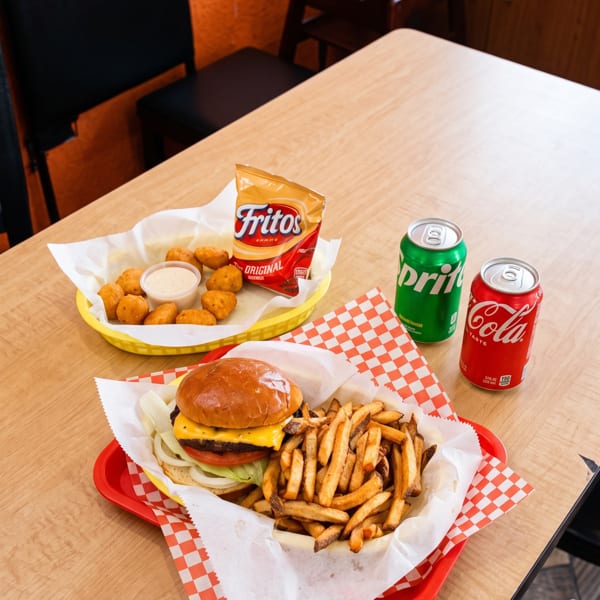 A burger and fries, chicken nuggets with Fritos, and two soda cans on a wooden table.