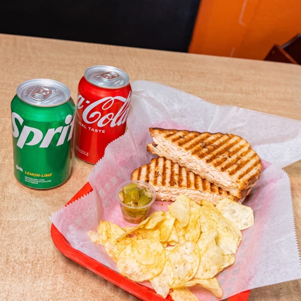 Grilled panini sandwich with chips and two soda cans on a table.