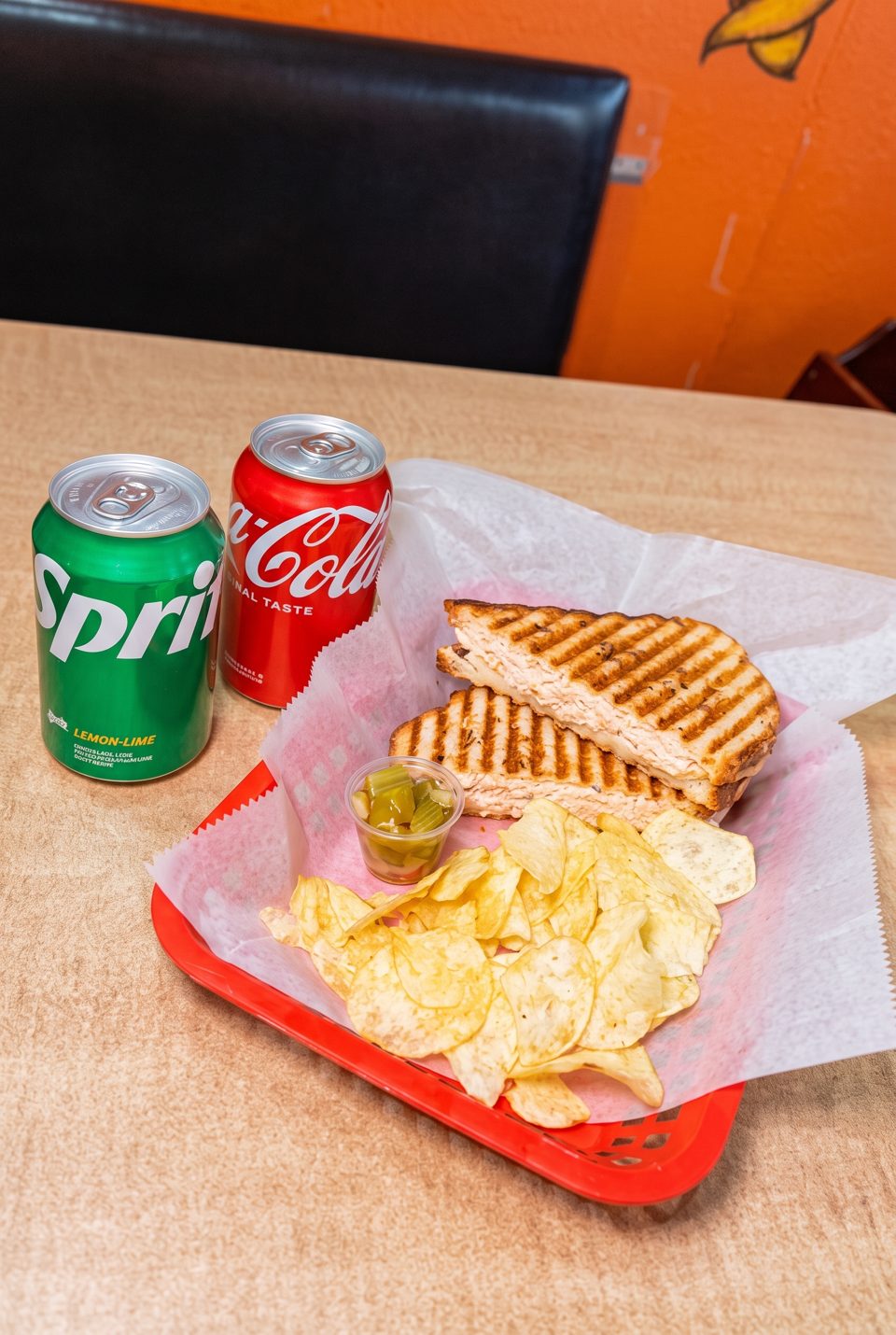 Grilled panini sandwich with chips and two soda cans on a table.