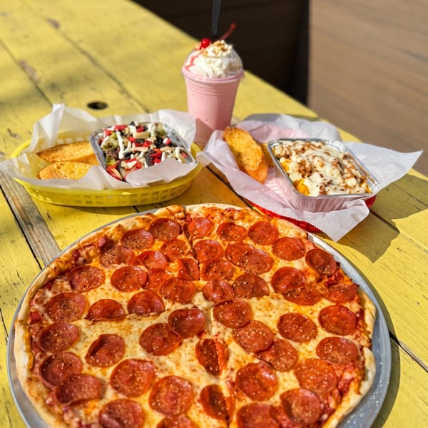 Pepperoni pizza, garlic bread, loaded fries, and a strawberry milkshake on a yellow outdoor table.