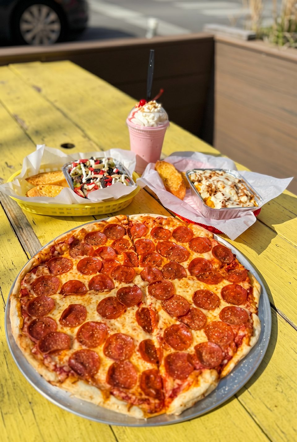 Pepperoni pizza, garlic bread, loaded fries, and a strawberry milkshake on a yellow outdoor table.