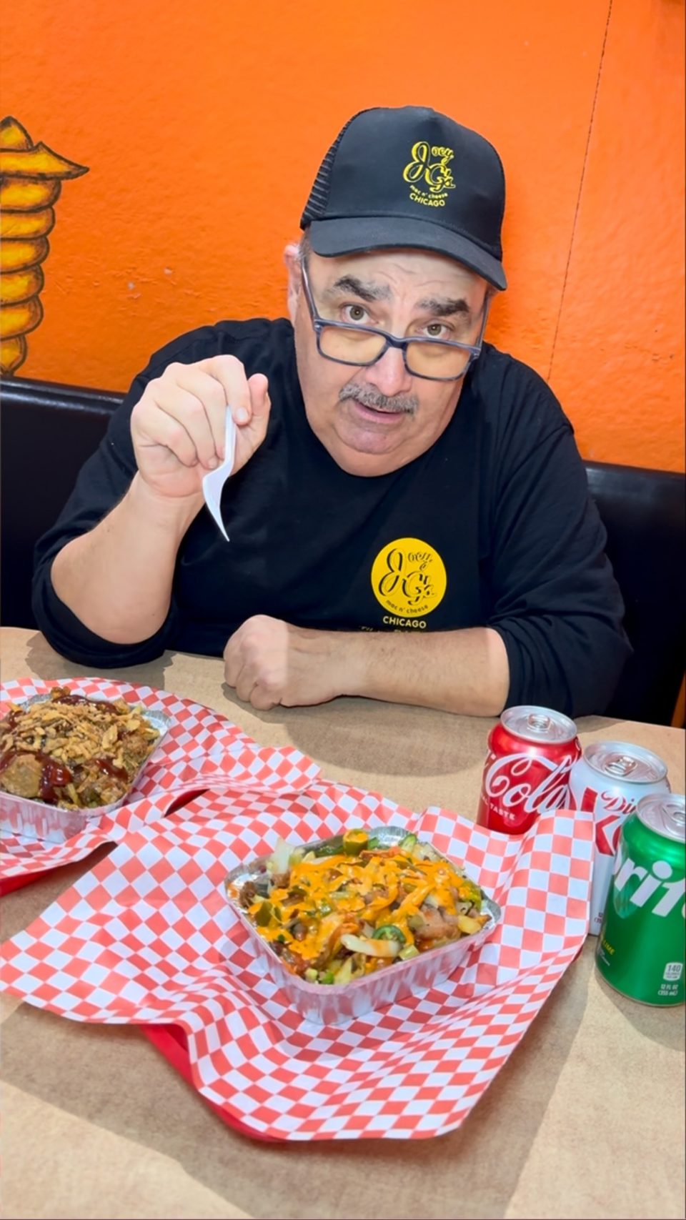 Man in a black hat and shirt with a yellow logo, holding a fork over two food trays.